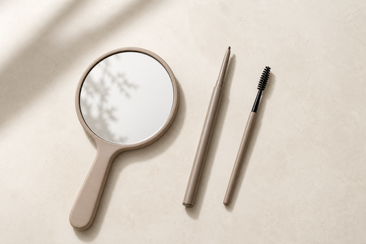 Four brow shape strokes drawn on a card beside a spoolie brush and hand mirror