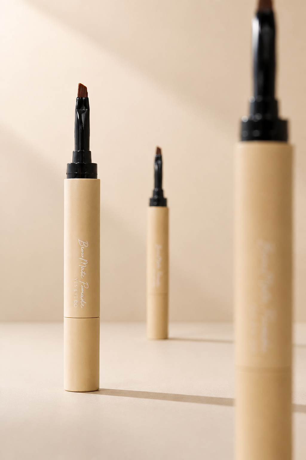 Three beige brow pomade pens standing on a warm beige background